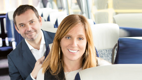 eine Dame und ein Herr lächeln in die Kamera und sitzen dabei hintereinander in einem Reisebus mit blauen Sitzen