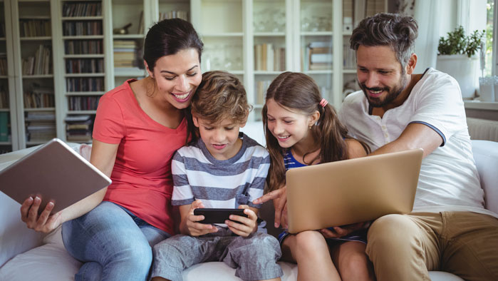 Familie, die auf einer Couch sitzt und auf mehreren Geräten gleichzeitig im Internet surft
